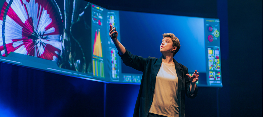 Een vrouw houdt een lezing op een podium. Ze wijst naar schermen achter haar waar verschillende soorten diagrammen op te zien zijn.