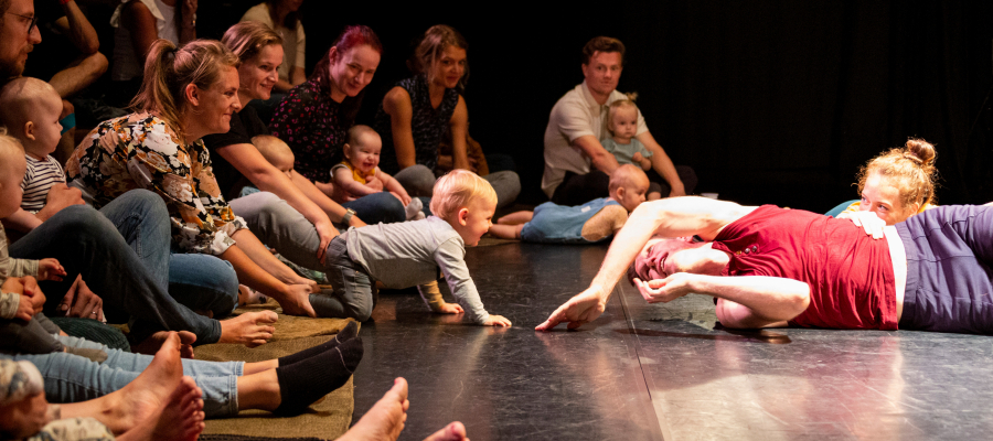 ouders met baby's en peuters zitten en liggen samen met twee artiesten van een theatergezelschap op een zwarte houten vloer