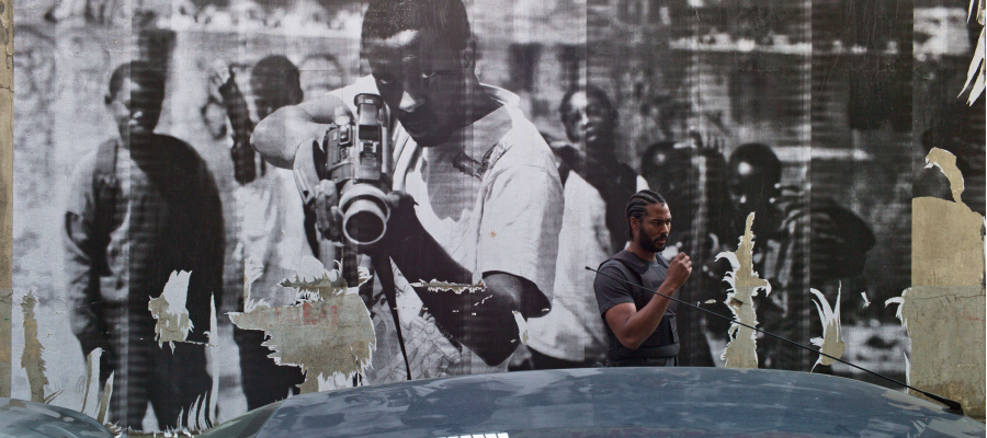 beeld uit de film: man staat achter een auto, geleund tegen een muur met een grote foto