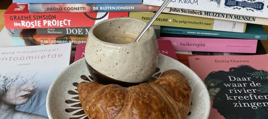 temidden van stapels boeken staat een kop koffie op een schoteltje met een croissant