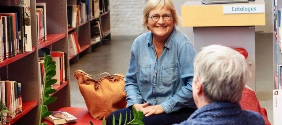 twee personen, een man en een vrouw, zitten tegenover elkaar in een bibliotheek en zijn in gesprek