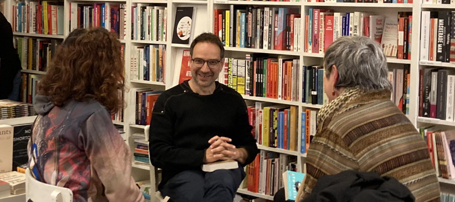 Drie personen zitten bij elkaar en zijn in gesprek. Op de achtergrond staat een grote gevulde boekenkast.