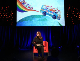 Fleur Pierets zit op een podium in een rode zetel met achter haar op een scherm een prent uit haar boek Love is Love, getekend door Fatinha Ramos.