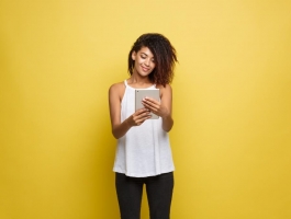 jonge vrouw staat voor een gele achtergrond en kijkt lachend naar haar tablet